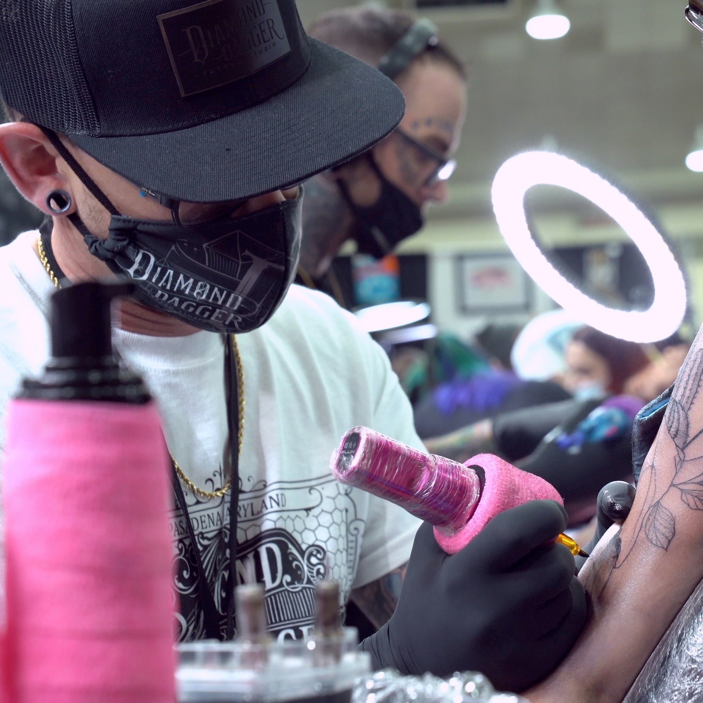 Tattoo artist at convention tattooing a client with a Bubblegum pink Flux wireless tattoo machine. Your Ultimate Tattoo Convention Guide