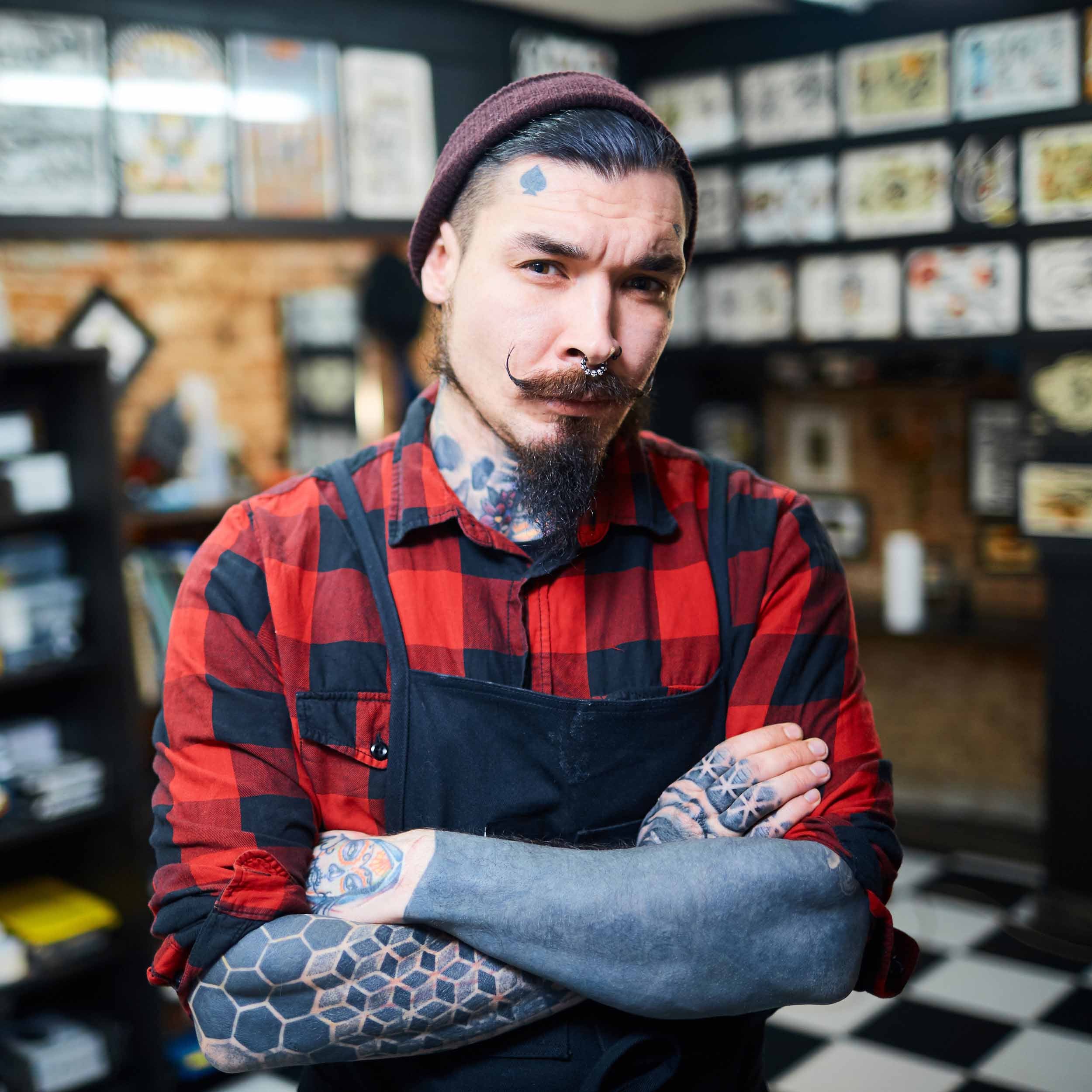 Piercer with tattoos looking skeptical with his arms folded TOP 10 Piercing Myths BUSTED - Don't Let Your Clients Be Fooled