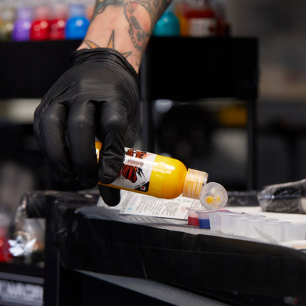 Gloved artist pouring yellow tattoo ink into a cap at a workstation.