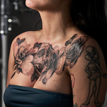 A woman showcases a large, detailed black and grey chest tattoo featuring a skull surrounded by flowers.