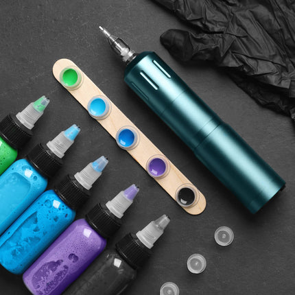 Clean flat lay of a teal rotary tattoo pen machine, matching vibrant ink bottles, ink caps on a wooden stick, and a black nitrile glove over a dark slate background.