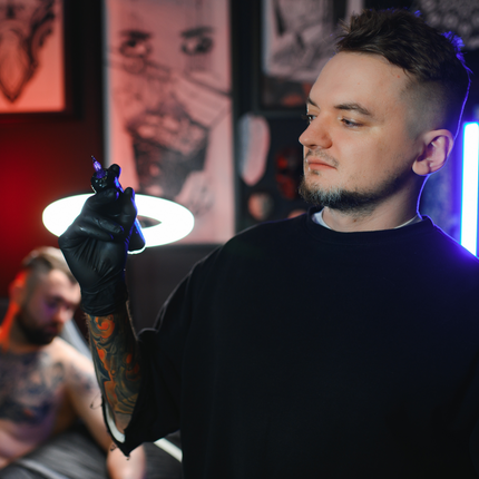A male tattoo artist in a black shirt looking at a tattoo needle cartridge in a studio setting.