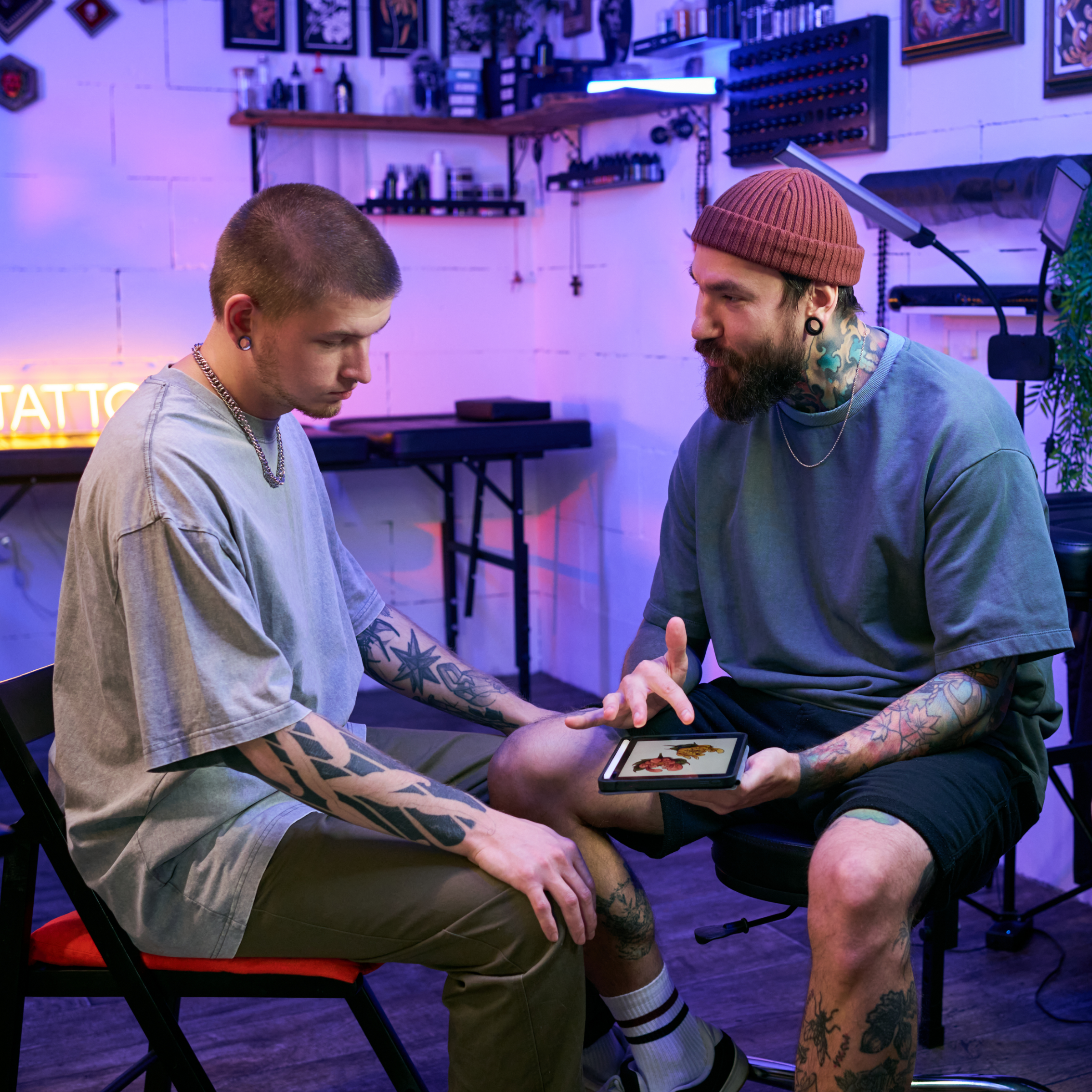 A tattoo artist and client discussing a design on a tablet in a neon-lit studio. Nobody Tells You Clients Don’t Remember Technique—They Remember Feelings