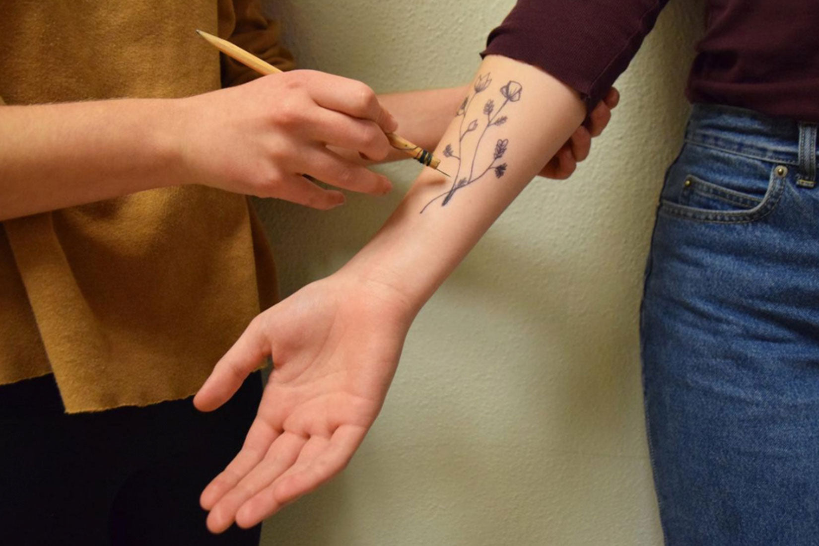 artist holding a stick and poke needle to an outstretched arm with floral tattoo design Hand Poked Tattoos vs. Tattoo Machine Tattoos